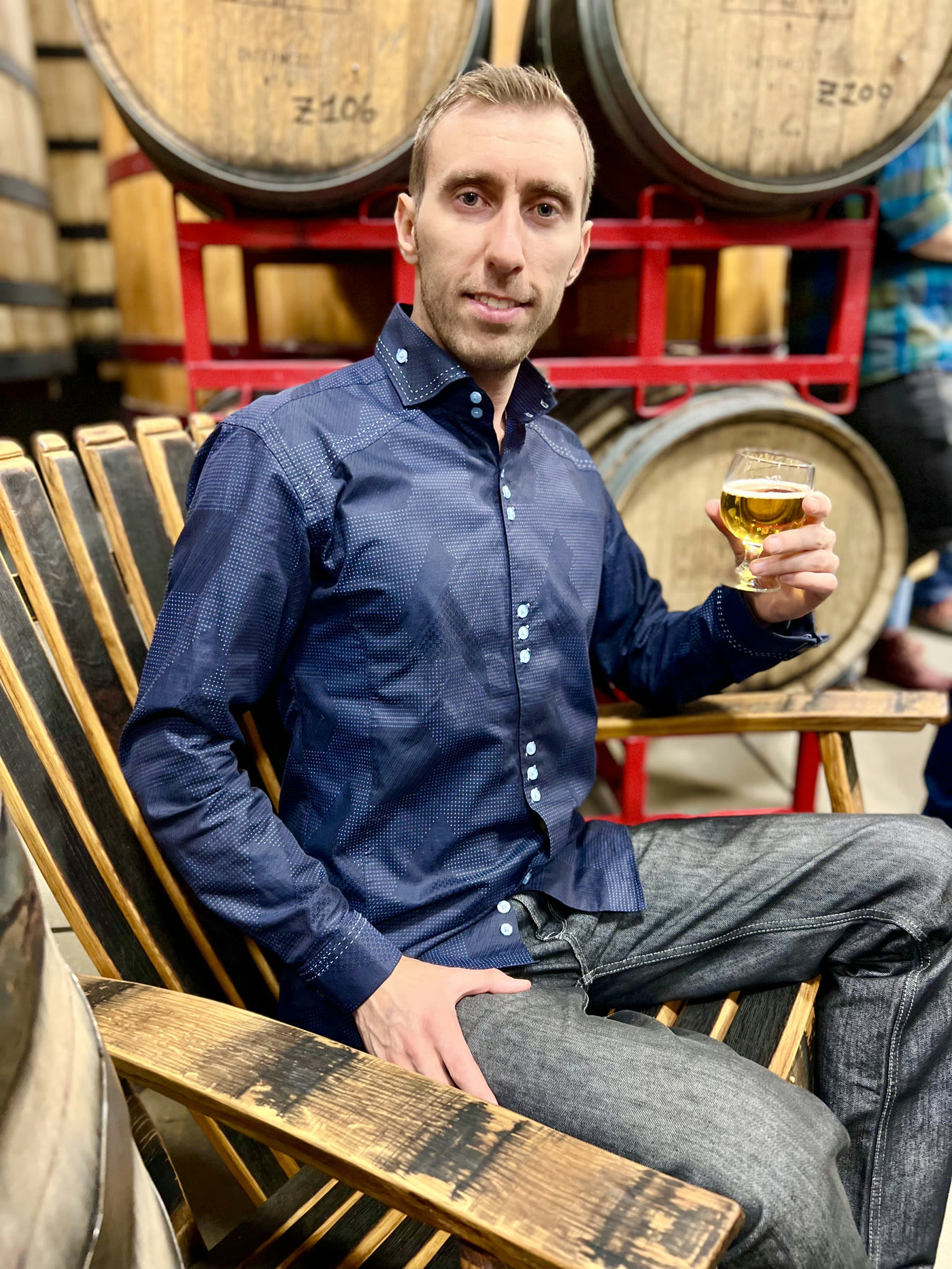 Pavel wearing a patterned navy shirt and gray jeans, seated beside wooden barrels